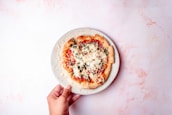 A hand holding a plate with a homemade pizza topped with cheese, tomato sauce, and vegetables. The background is a light marbled surface, creating a soft contrast with the vibrant colors of the pizza.