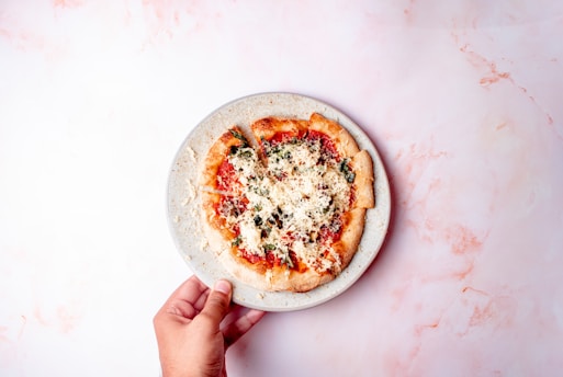 A hand holding a plate with a homemade pizza topped with cheese, tomato sauce, and vegetables. The background is a light marbled surface, creating a soft contrast with the vibrant colors of the pizza.