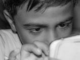 A child intensely focused on a device held closely in their hands, with fingers visible on the device. The close-up shot highlights their curious and engaged expression, emphasizing their eyes and eyelashes.