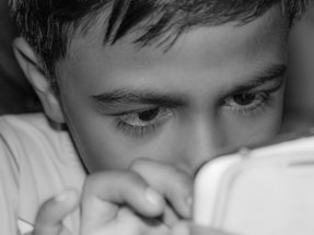 A child intensely focused on a device held closely in their hands, with fingers visible on the device. The close-up shot highlights their curious and engaged expression, emphasizing their eyes and eyelashes.