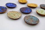 A collection of small, colorful ceramic dishes with intricate patterns scattered across a light, neutral background. The dishes feature various designs, including swirling lines and geometric shapes, in vibrant shades of blue, green, and yellow.
