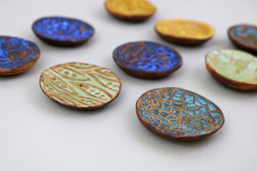 A collection of small, colorful ceramic dishes with intricate patterns scattered across a light, neutral background. The dishes feature various designs, including swirling lines and geometric shapes, in vibrant shades of blue, green, and yellow.