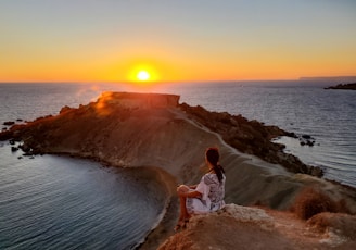 woman in white shirt standing on brown rock near body of water during sunset