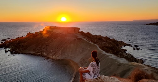 woman in white shirt standing on brown rock near body of water during sunset