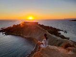 woman in white shirt standing on brown rock near body of water during sunset