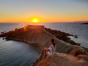 woman in white shirt standing on brown rock near body of water during sunset