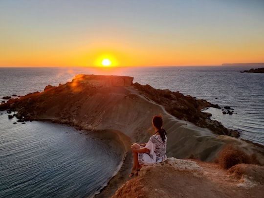 woman in white shirt standing on brown rock near body of water during sunset in Għajn Tuffieħa Malta