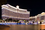 A large, brightly lit hotel complex is situated along a waterfront, with ornate architecture and numerous windows illuminated at night. The scene includes a reflective water body in the foreground and a clear dark sky above.