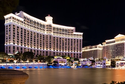 A large, brightly lit hotel complex is situated along a waterfront, with ornate architecture and numerous windows illuminated at night. The scene includes a reflective water body in the foreground and a clear dark sky above.