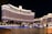 A large, brightly lit hotel complex is situated along a waterfront, with ornate architecture and numerous windows illuminated at night. The scene includes a reflective water body in the foreground and a clear dark sky above.