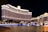 A large, brightly lit hotel complex is situated along a waterfront, with ornate architecture and numerous windows illuminated at night. The scene includes a reflective water body in the foreground and a clear dark sky above.