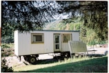 A mobile structure resembling a trailer surrounded by lush greenery and trees. The structure has a door and windows with barred coverings. Nearby, there is an unmarked wooden sign leaning against the trailer.