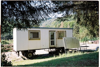 A mobile structure resembling a trailer surrounded by lush greenery and trees. The structure has a door and windows with barred coverings. Nearby, there is an unmarked wooden sign leaning against the trailer.