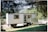 A mobile structure resembling a trailer surrounded by lush greenery and trees. The structure has a door and windows with barred coverings. Nearby, there is an unmarked wooden sign leaning against the trailer.