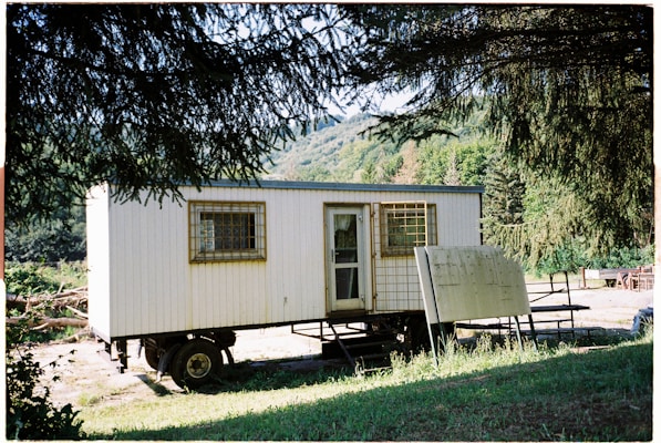 A mobile structure resembling a trailer surrounded by lush greenery and trees. The structure has a door and windows with barred coverings. Nearby, there is an unmarked wooden sign leaning against the trailer.