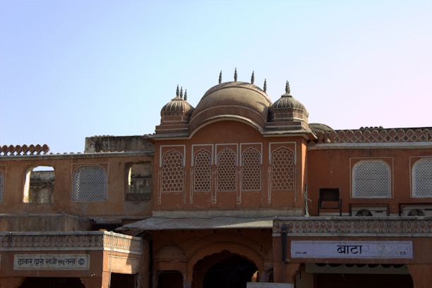 An architectural structure with intricate lattice windows and domed rooftops, displaying a traditional design with earthy tones. The facade features ornate detailing and a symmetrical layout, with Hindi signs visible at the bottom.