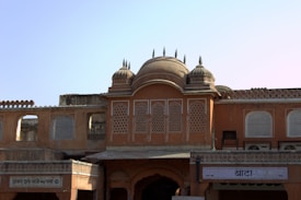 An architectural structure with intricate lattice windows and domed rooftops, displaying a traditional design with earthy tones. The facade features ornate detailing and a symmetrical layout, with Hindi signs visible at the bottom.