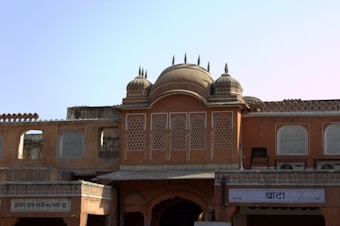 An architectural structure with intricate lattice windows and domed rooftops, displaying a traditional design with earthy tones. The facade features ornate detailing and a symmetrical layout, with Hindi signs visible at the bottom.