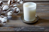 A white candle in a frosted glass container labeled 'shore soap co.' sits on a decorative metal tray. To the left, cotton branches with fluffy white cotton bolls rest on a rustic wooden surface.