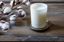 A white candle in a frosted glass container labeled 'shore soap co.' sits on a decorative metal tray. To the left, cotton branches with fluffy white cotton bolls rest on a rustic wooden surface.