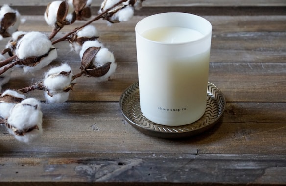 A white candle in a frosted glass container labeled 'shore soap co.' sits on a decorative metal tray. To the left, cotton branches with fluffy white cotton bolls rest on a rustic wooden surface.