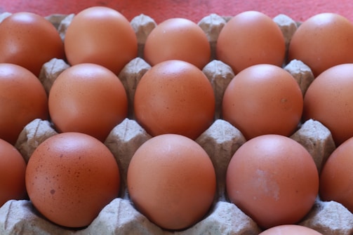 A carton containing multiple brown eggs arranged in a grid pattern. The eggs are uniformly placed in individual slots of the carton, which is made of a light, textured material. The background is a red or pinkish fabric.