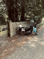 A motorcycle is parked beside a stone wall along a quiet road. The bike has a blue and black color scheme with a windshield and a side bag attached. A white helmet is resting on the seat. The background includes dense green foliage and a wooden gate.