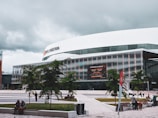 A modern building with a large sign that reads 'Centre Vidéotron' is prominently positioned in a plaza. The structure has a sleek design with glass windows and metallic panels. Trees and people are scattered throughout the paved courtyard, and a digital billboard displays information about an event. The sky above is overcast with gray clouds.