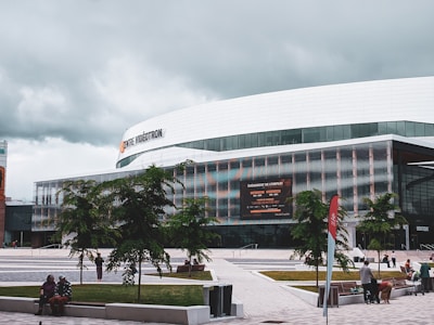 A modern building with a large sign that reads 'Centre Vidéotron' is prominently positioned in a plaza. The structure has a sleek design with glass windows and metallic panels. Trees and people are scattered throughout the paved courtyard, and a digital billboard displays information about an event. The sky above is overcast with gray clouds.