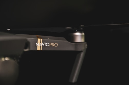 A close-up view of a drone with the branding 'Mavic Pro' visible on its arm. The device appears sleek and modern with a matte finish. Lighting is low, highlighting the drone's contours and design.