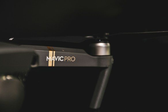 A close-up view of a drone with the branding 'Mavic Pro' visible on its arm. The device appears sleek and modern with a matte finish. Lighting is low, highlighting the drone's contours and design.
