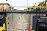 A large industrial lock chamber is depicted, with concrete walls and a dry, empty basin. Scaffolding is erected along the walls, suggesting maintenance or construction work. Two people wearing safety gear are visible, one near the middle and another further inside the lock.