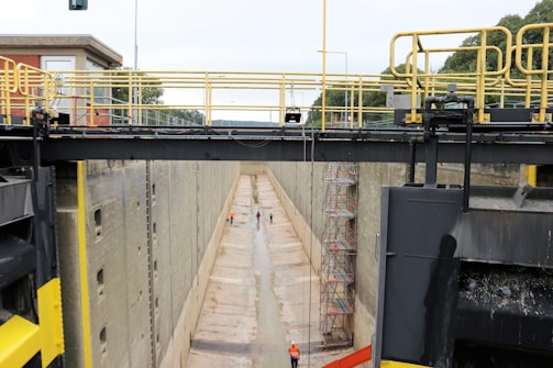 A large industrial lock chamber is depicted, with concrete walls and a dry, empty basin. Scaffolding is erected along the walls, suggesting maintenance or construction work. Two people wearing safety gear are visible, one near the middle and another further inside the lock.