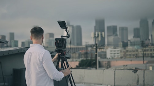 A person in a white shirt is operating a video camera on a tripod, positioned on a rooftop with a cityscape view in the background. The skyline appears hazy and overcast, suggesting cloudy weather.