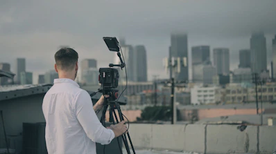 A person in a white shirt is operating a video camera on a tripod, positioned on a rooftop with a cityscape view in the background. The skyline appears hazy and overcast, suggesting cloudy weather.