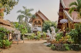 A picturesque courtyard is surrounded by traditional Thai architecture, featuring ornate carvings and roof designs. Lush greenery, including palm trees and various plants, adds to the serene ambiance. Decorative statues are placed throughout the area, and there is a sense of calm and spiritual tranquility.