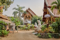 A picturesque courtyard is surrounded by traditional Thai architecture, featuring ornate carvings and roof designs. Lush greenery, including palm trees and various plants, adds to the serene ambiance. Decorative statues are placed throughout the area, and there is a sense of calm and spiritual tranquility.