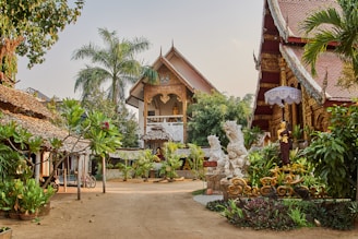 A picturesque courtyard is surrounded by traditional Thai architecture, featuring ornate carvings and roof designs. Lush greenery, including palm trees and various plants, adds to the serene ambiance. Decorative statues are placed throughout the area, and there is a sense of calm and spiritual tranquility.