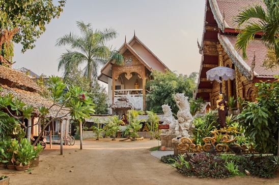 A picturesque courtyard is surrounded by traditional Thai architecture, featuring ornate carvings and roof designs. Lush greenery, including palm trees and various plants, adds to the serene ambiance. Decorative statues are placed throughout the area, and there is a sense of calm and spiritual tranquility.