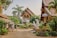 A picturesque courtyard is surrounded by traditional Thai architecture, featuring ornate carvings and roof designs. Lush greenery, including palm trees and various plants, adds to the serene ambiance. Decorative statues are placed throughout the area, and there is a sense of calm and spiritual tranquility.