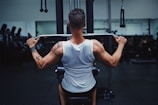 A person wearing a white tank top is seated on a gym machine, performing a pull-down exercise. They are focused on working out, with visible muscular back and tattoo on the left arm. The background has other gym equipment silhouetted in low lighting, giving a concentrated atmosphere.