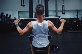 A person wearing a white tank top is seated on a gym machine, performing a pull-down exercise. They are focused on working out, with visible muscular back and tattoo on the left arm. The background has other gym equipment silhouetted in low lighting, giving a concentrated atmosphere.