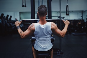 A person wearing a white tank top is seated on a gym machine, performing a pull-down exercise. They are focused on working out, with visible muscular back and tattoo on the left arm. The background has other gym equipment silhouetted in low lighting, giving a concentrated atmosphere.