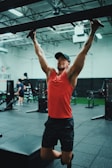 A person in a gym is performing pull-ups using a bar. They are wearing a red tank top, black shorts, and a cap. The gym has a variety of fitness equipment, with other people exercising in the background. The setting is spacious, featuring a high ceiling and industrial-style interior.