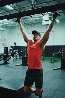 A person in a gym is performing pull-ups using a bar. They are wearing a red tank top, black shorts, and a cap. The gym has a variety of fitness equipment, with other people exercising in the background. The setting is spacious, featuring a high ceiling and industrial-style interior.