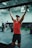 A person in a gym is performing pull-ups using a bar. They are wearing a red tank top, black shorts, and a cap. The gym has a variety of fitness equipment, with other people exercising in the background. The setting is spacious, featuring a high ceiling and industrial-style interior.
