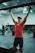 A person in a gym is performing pull-ups using a bar. They are wearing a red tank top, black shorts, and a cap. The gym has a variety of fitness equipment, with other people exercising in the background. The setting is spacious, featuring a high ceiling and industrial-style interior.