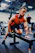 A smiling gym member lifting weights with orange and dark-themed gym equipment in the background.