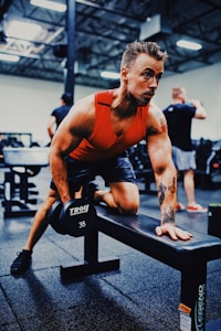 A muscular individual is engaged in a weightlifting exercise at a gym, using a dumbbell while leaning on a bench. They are wearing a sleeveless orange top and exhibit a focused expression. The gym background features other fitness enthusiasts and various equipment under bright artificial lighting.