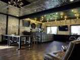 A modern cafe interior with industrial-style decor. The space features wooden flooring and a high ceiling with geometric lighting fixtures. A long counter with a series of small drawers runs alongside one wall, topped with a menu board and coffee-making equipment. Seating includes leather chairs and a long communal table with benches. Exposed brick walls add texture, while large windows provide natural light.